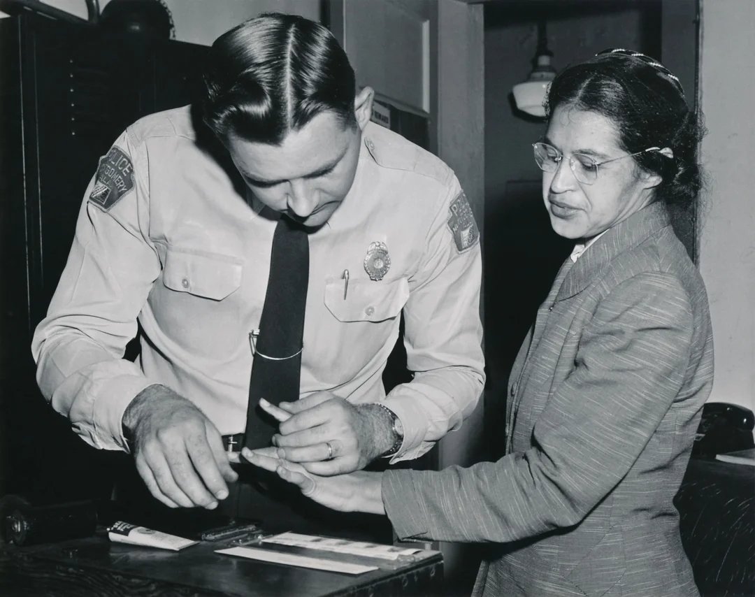 A police officer in uniform takes the fingerprints of a woman wearing glasses and a suit jacket in an indoor setting. The woman looks down at her hand as the officer presses her finger onto a card.