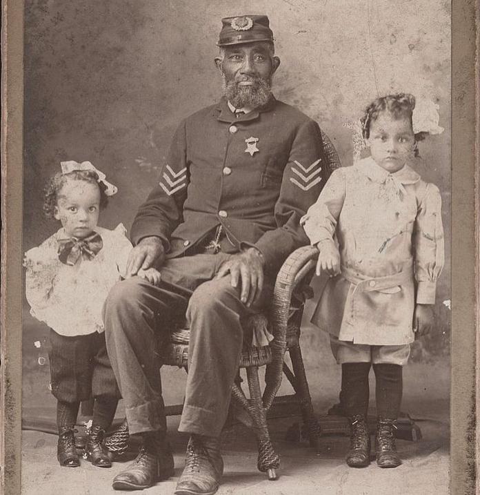 A bearded Black man in a decorated military uniform sits on a chair, flanked by two young children standing on either side, both wearing bows and formal clothing, in an old sepia-toned photograph.
