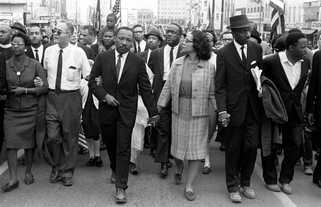 Black-and-white photo of civil rights activists, including Martin Luther King Jr., marching arm in arm down a city street with a diverse group of men and women, some holding hands and American flags visible in the background.