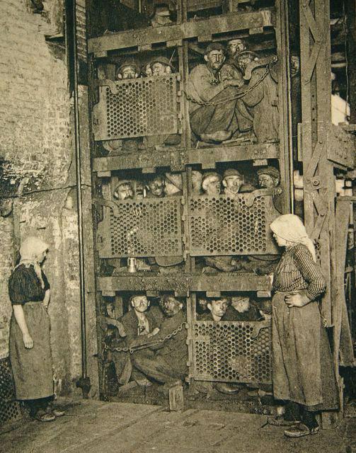 Historic black and white photo showing coal miners crowded into a large metal elevator cage, preparing to descend into a mine shaft, while two women in work clothes stand nearby watching.