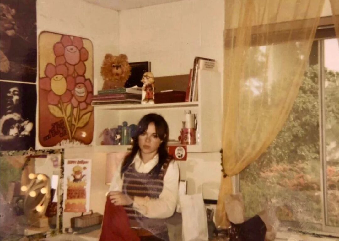 A young woman stands in a vintage room by a cluttered desk, holding a red cloth. The room has posters, shelves with books and toys, a window with yellow curtains, and sunlight streaming in. The overall tone is nostalgic.