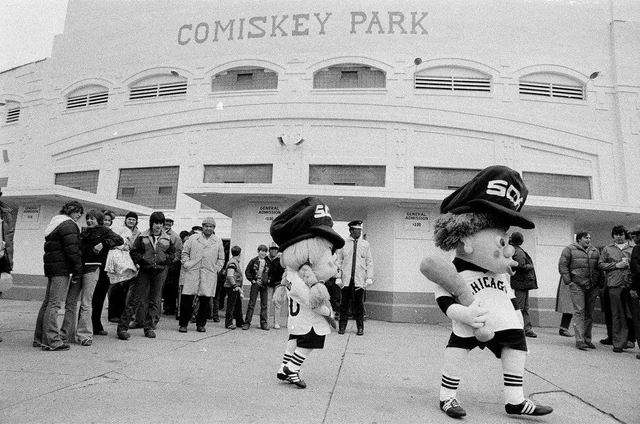 Black-and-white photo of people lined up outside Comiskey Park. Two people in large Chicago White Sox mascot costumes walk in front, wearing baseball uniforms and oversized hats. The stadium entrance is visible in the background.