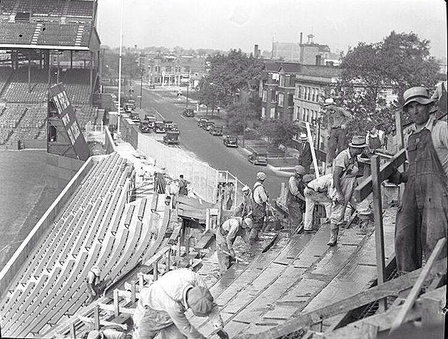 Workers construct or repair stadium seating, using tools and materials, while wearing work clothes and hats. The scene overlooks a city street with cars and buildings in the background.
