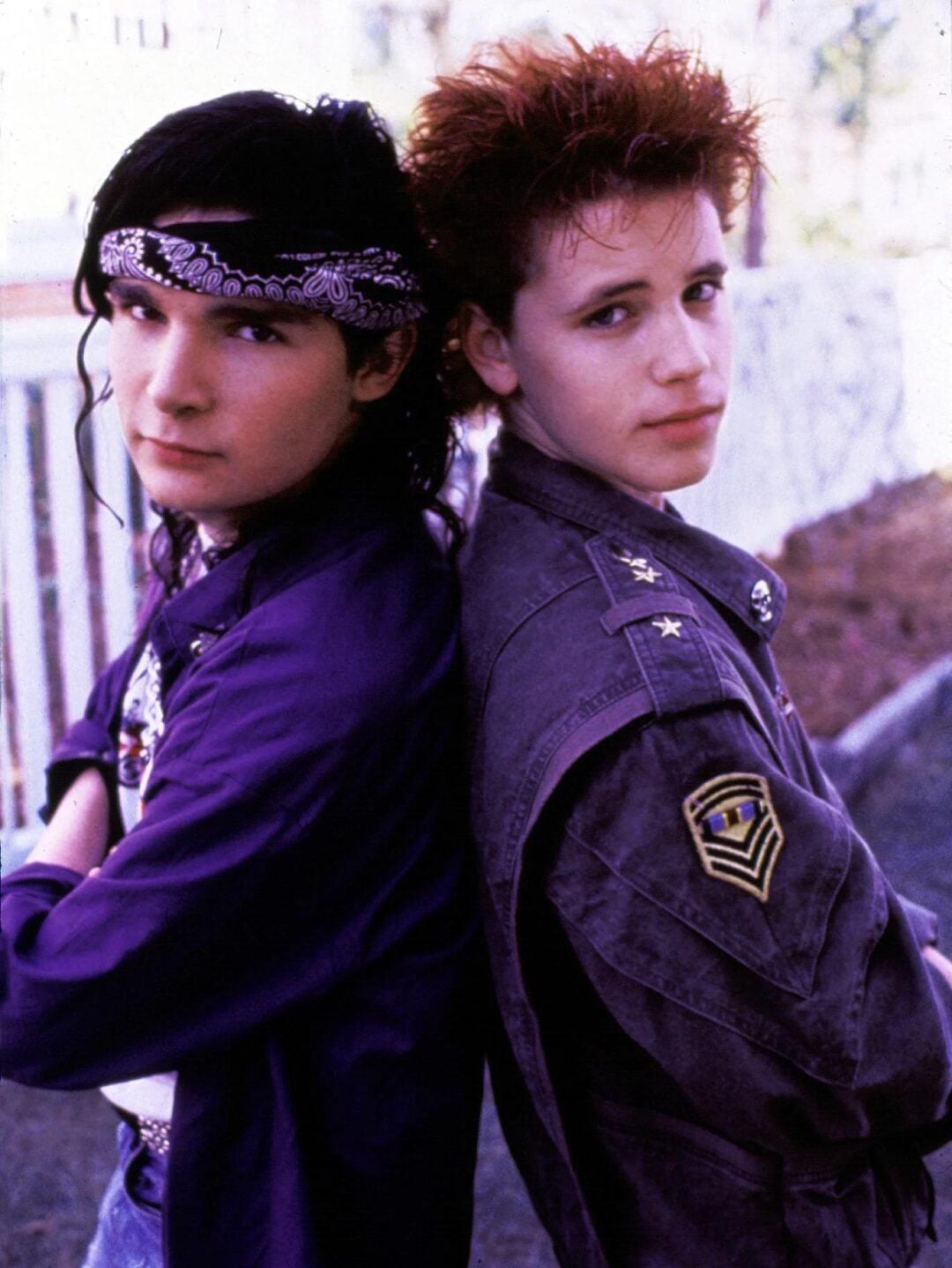 Two teenage boys stand back to back outdoors. One has long, dark hair with a headband and a purple shirt; the other has short, spiky red hair and wears a military-style jacket with patches. Both look at the camera.
