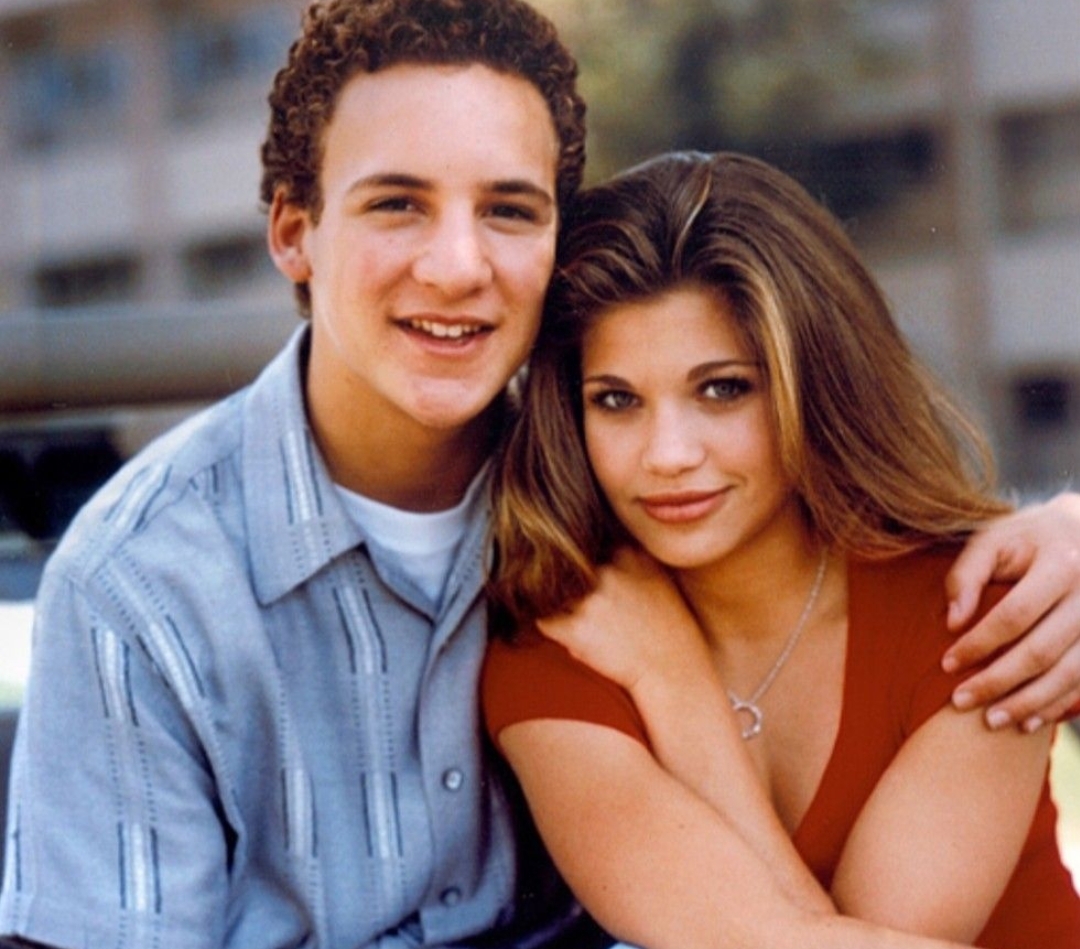 A teenage boy with short curly hair and a striped shirt sits outside with his arm around a teenage girl with long straight hair and a red top. They both smile at the camera.