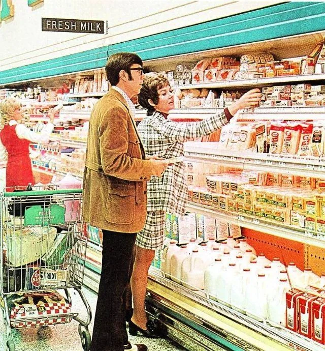 A man and woman shop for dairy products in a supermarket aisle, standing near shelves of milk, cheese, and other products. A woman in red pushes a cart in the background. The scene appears to be from the 1970s.