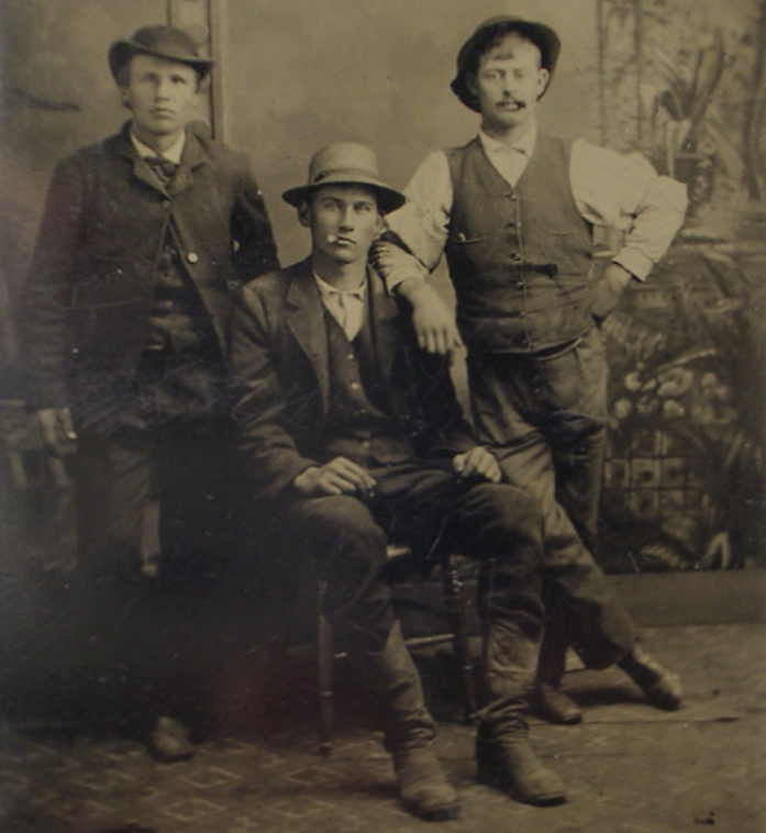 Three men in old-fashioned suits and hats pose for a vintage photograph. Two stand while one sits on a chair. The background has a painted, rustic scene. All three wear serious expressions.