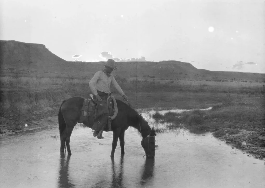 A cowboy on horseback leans forward as his horse drinks from a shallow stream, with open plains and distant mesas in the background under a bright sky.