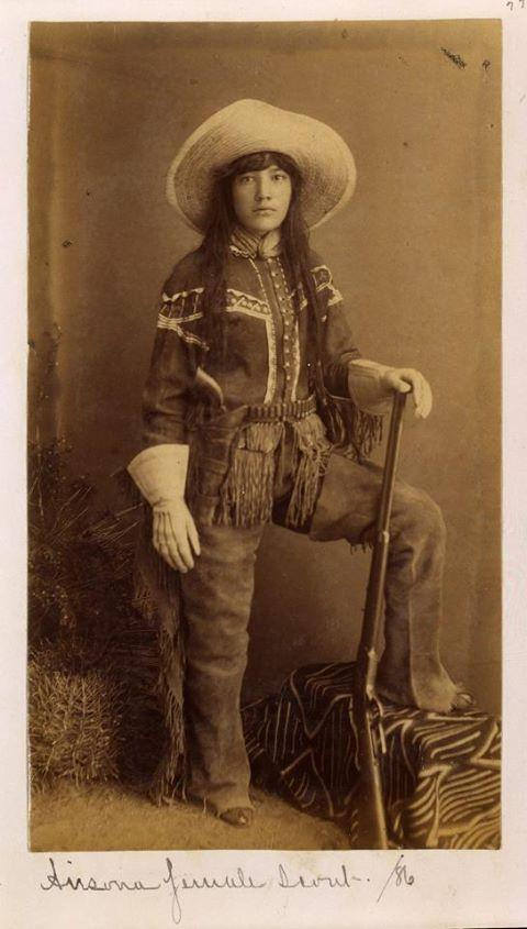 A young woman in a fringed western outfit and wide-brimmed hat poses with one knee on a draped object, holding a rifle. Handwritten text below reads, "Lissena Female Scout.