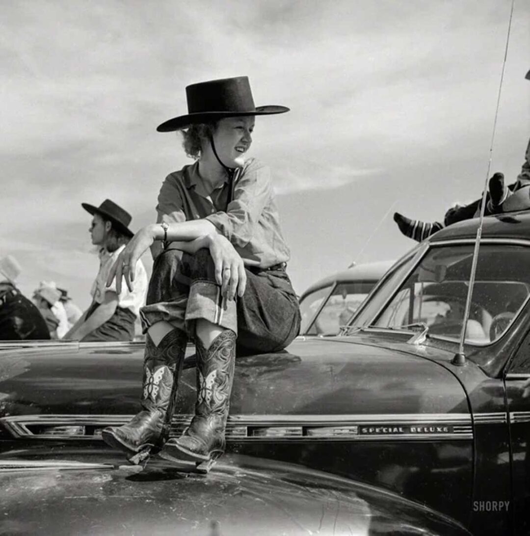 A woman in a cowboy hat and boots sits on the hood of a vintage car, smiling and looking to the side. Other people in western attire and cars are visible in the background under a partly cloudy sky.