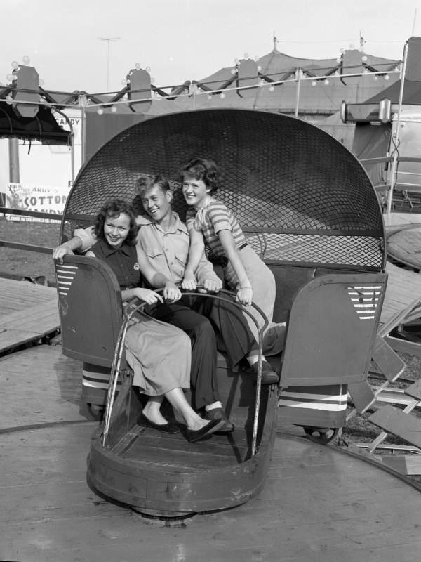 Three smiling young adults sit together in a spinning amusement park ride, likely at a fairground, with tents and a sign in the background. Two women flank a man; all appear happy and excited.
