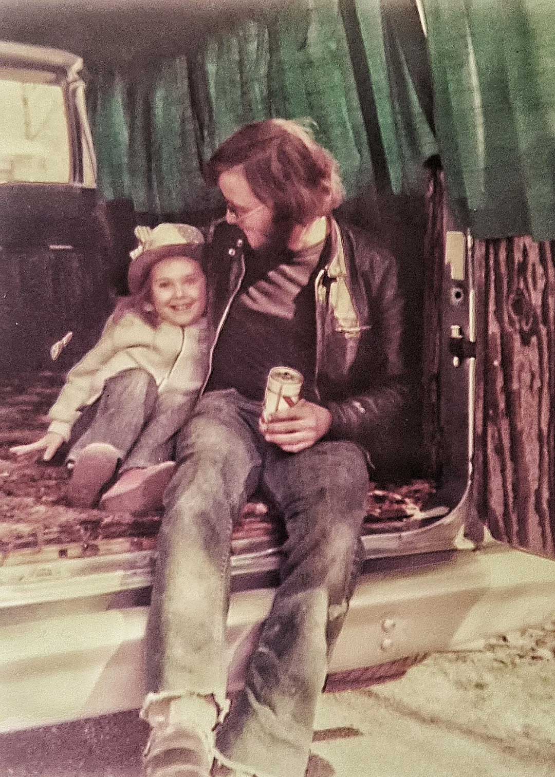 A man and a young girl sit together in the open back of a van with green curtains. The man holds a can and looks at the girl, who is smiling and wearing a hat. Both appear relaxed and happy.