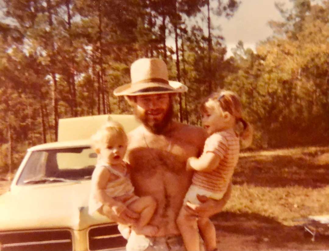 A bearded man wearing a straw hat stands shirtless outdoors, holding two young children in his arms. A yellow car and trees are visible in the background under sunny skies.