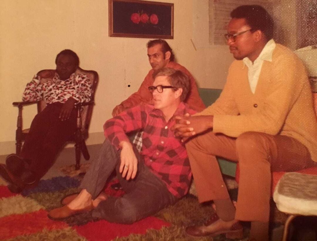 Four men watch something off camera in a living room. One sits in a chair, two sit on the floor, and one sits on a couch. The room has a colorful rug, framed wall art, and a window with blinds.