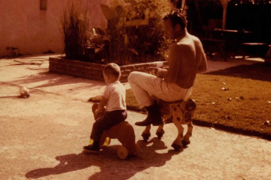 A man and a young child sit on toy animals in a sunlit backyard, both facing away. The scene is relaxed, with long shadows stretching across the concrete patio and grass in the background.