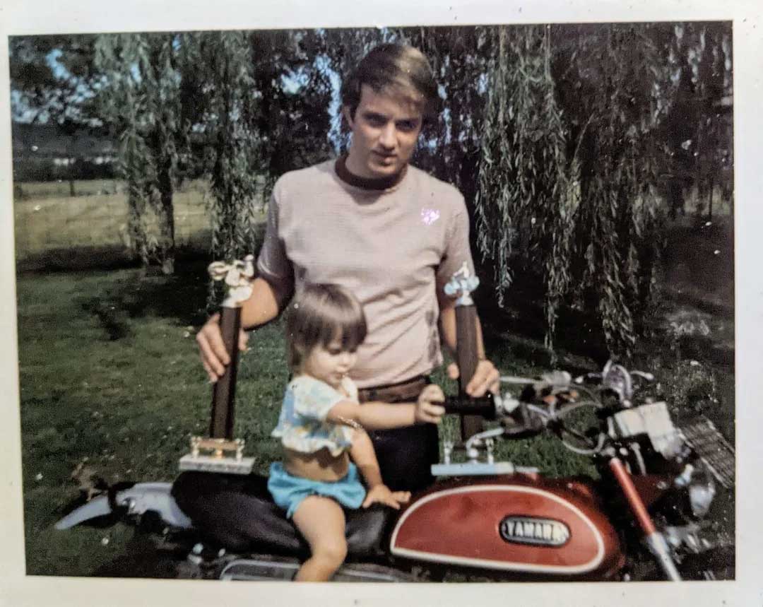 A young man stands outdoors holding two trophies, while a small child sits on a red Yamaha motorcycle in front of him. A tree and grassy field are visible in the background.