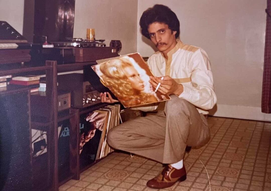 A man with dark hair and a mustache squats by a record player, holding a vinyl album with a woman's portrait on the cover. He wears a light shirt and brown pants, surrounded by records on a patterned floor.