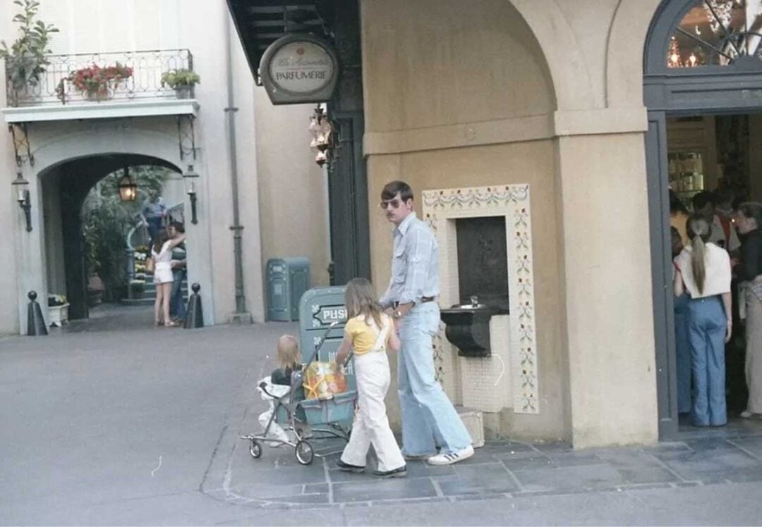 A man in sunglasses stands with two young children, one pushing a stroller, near a tiled drinking fountain and a trash can under an arched building in a quaint outdoor setting. Other people are visible in the background.