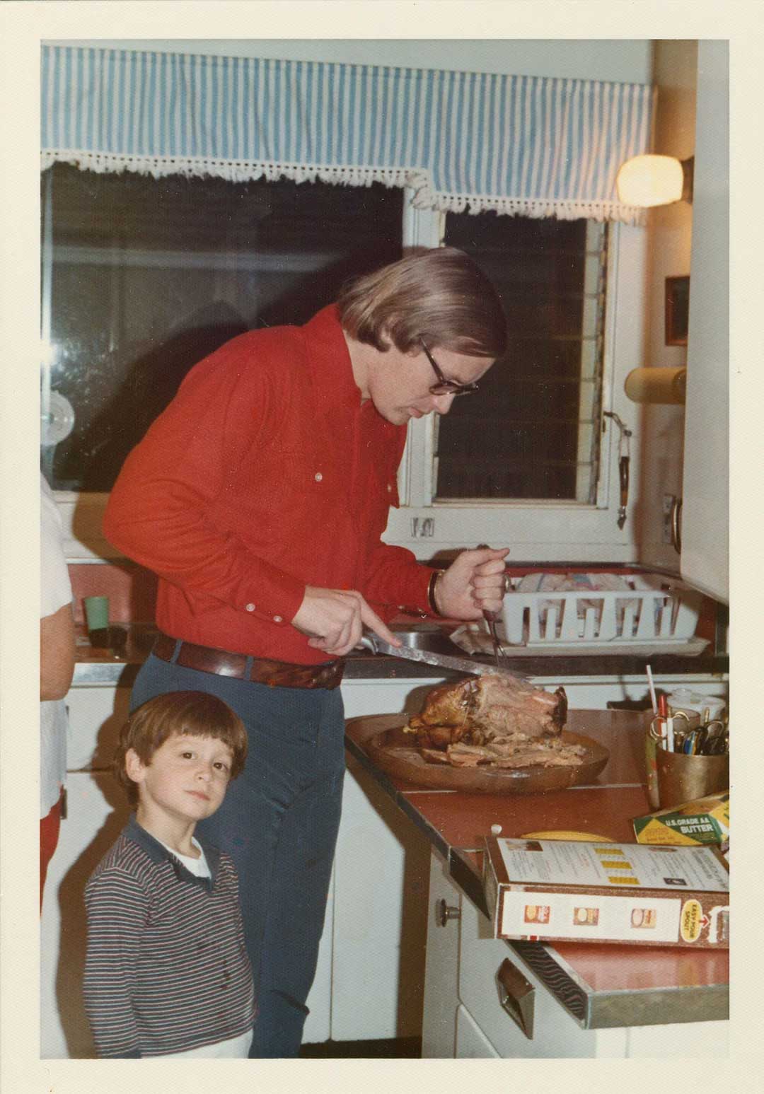A man in a red shirt slices a roast turkey on a kitchen counter while a young boy in a striped shirt stands nearby, looking at the camera. The kitchen has white cabinets and a window with a blue striped valance.