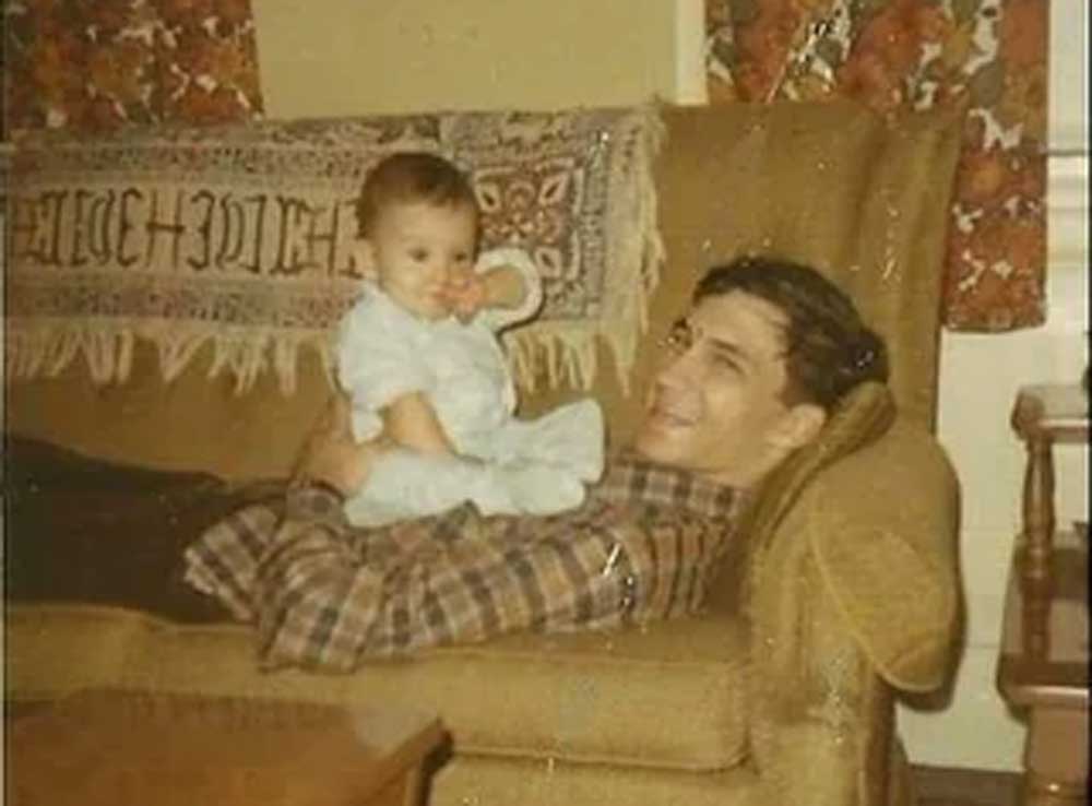 A man in a plaid shirt lies on a brown couch, smiling and holding a baby dressed in light blue pajamas. They appear happy and relaxed in a cozy room with patterned curtains and a decorative blanket.