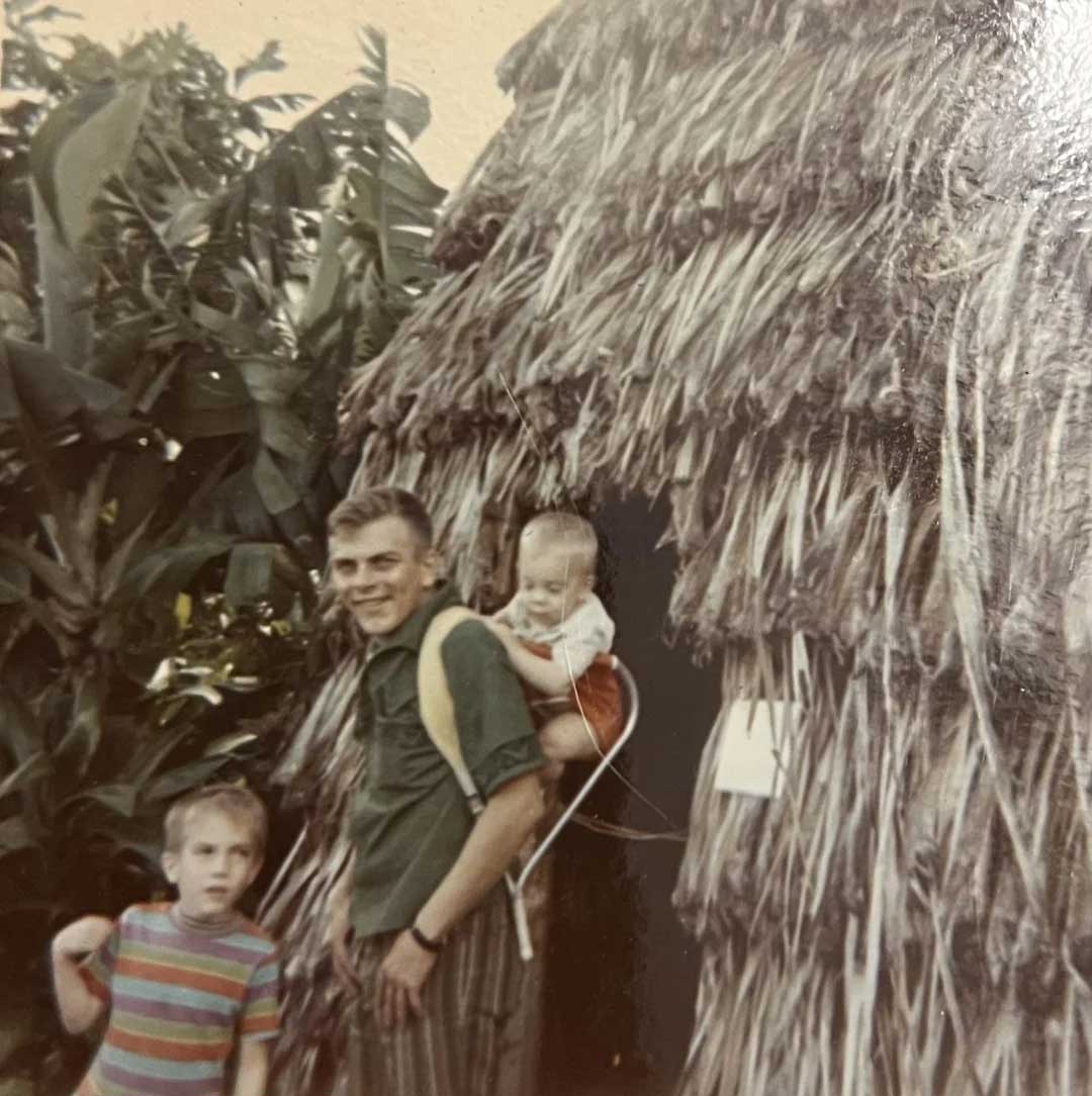 A man with two children stands outside a hut made of dried leaves. One child is on his back in a carrier, and another stands nearby holding a bag. Lush green plants are visible in the background. The photo appears vintage.