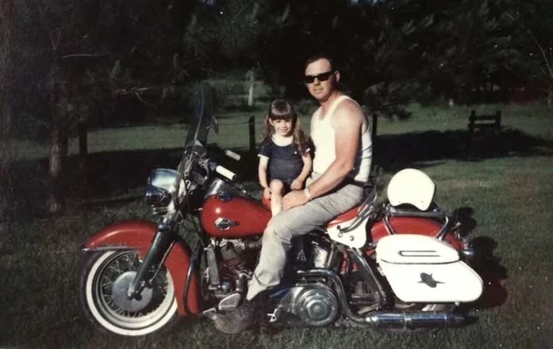 A man in sunglasses and a sleeveless shirt sits on a red and white motorcycle with a young girl in a blue dress sitting in front of him. They are outdoors on grass with trees in the background.