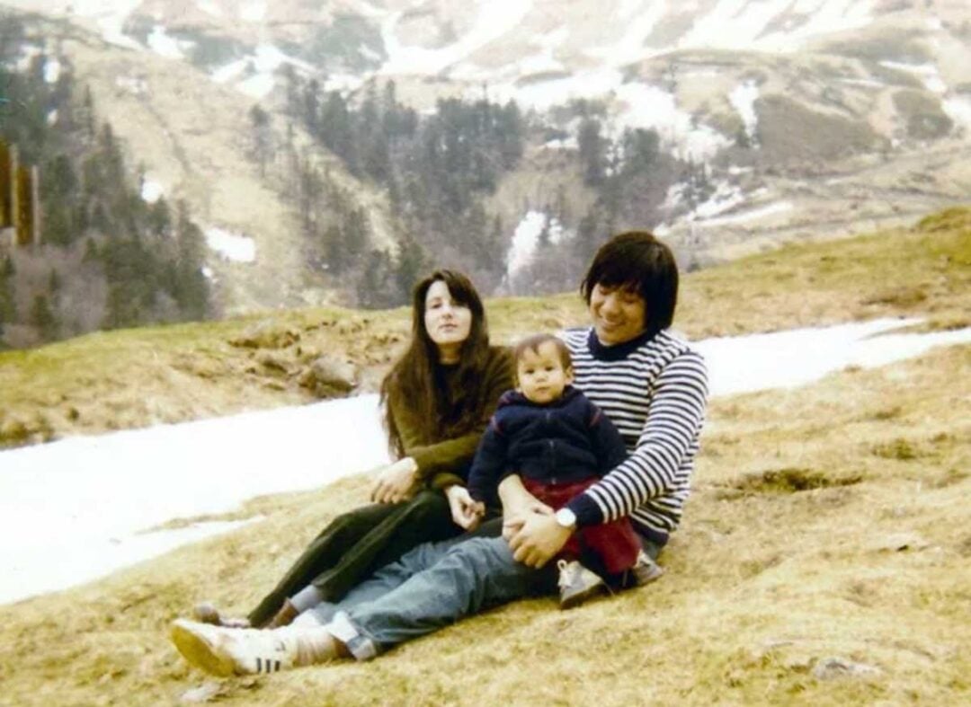 A man, woman, and young child sit on dry grass in front of a mountainous landscape with patches of snow. The man wears a striped shirt, the woman has long hair, and the child sits on the man's lap.