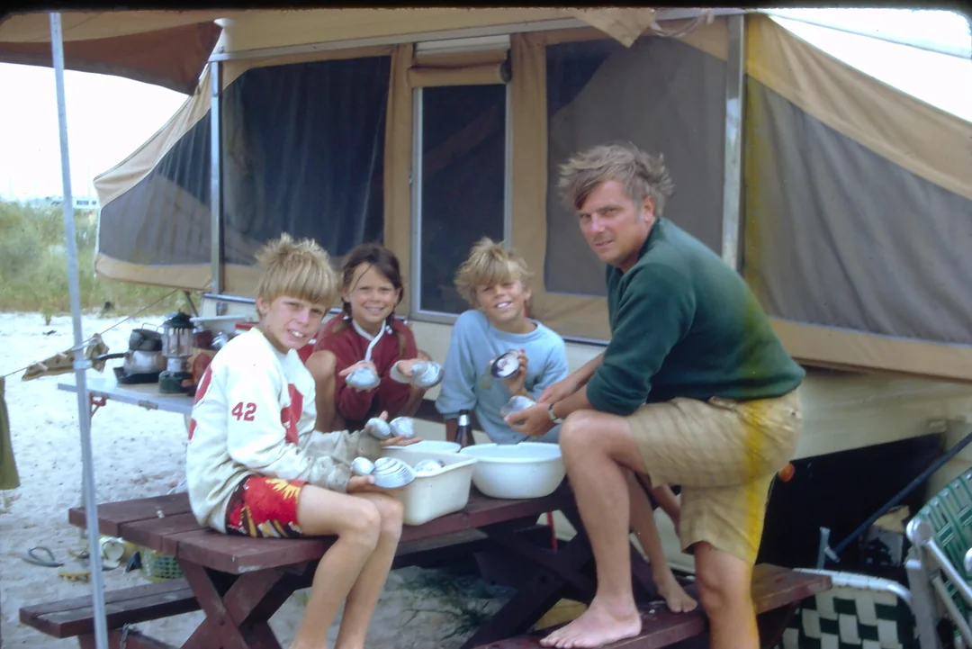 Three children and a man sit at a picnic table beside a camper tent, smiling and holding bowls of food. Camping gear is visible around them, and they appear to be enjoying a meal outdoors.