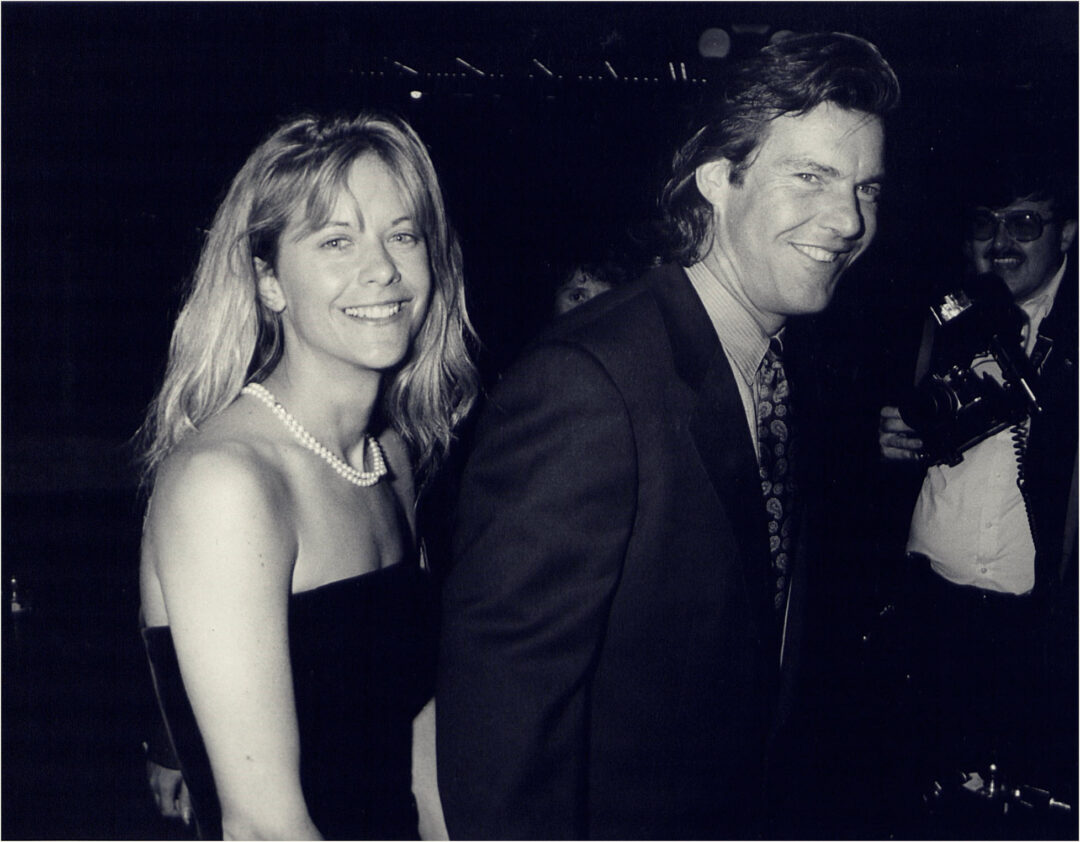 A smiling woman in a strapless dress and pearl necklace stands next to a man in a suit and tie at an event, with a photographer and another person visible in the background.