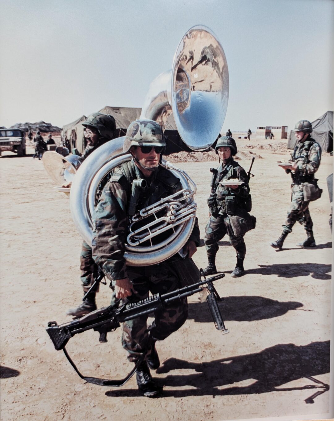 A soldier in camouflage gear carries a sousaphone and a rifle in a desert camp, with other armed soldiers and tents visible in the background under a clear sky.