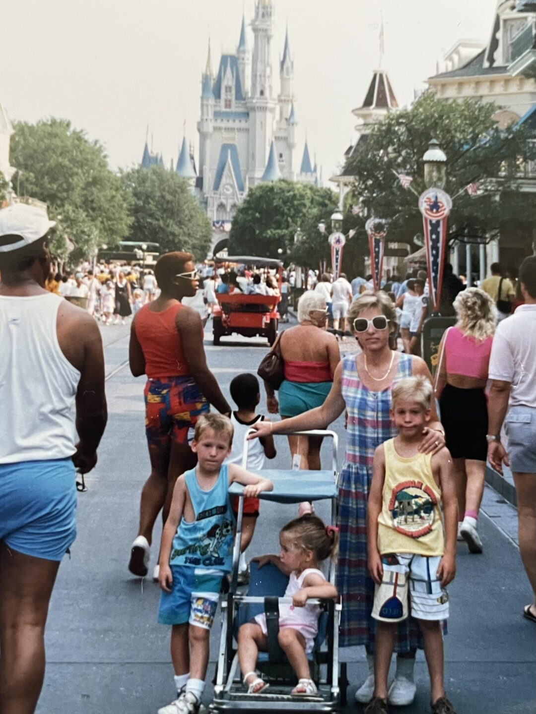 A woman and three children, one in a stroller, pose on a busy street at a theme park with a large castle in the background. People walk around them on a sunny day.