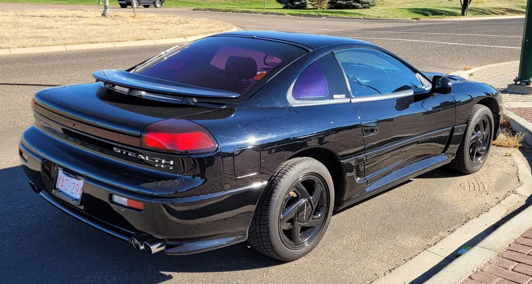 A black Mitsubishi Stealth sports car is parked on the side of a street, featuring tinted windows, a rear spoiler, and black rims. The image is taken from the rear-left, showing the car's sleek, aerodynamic design.