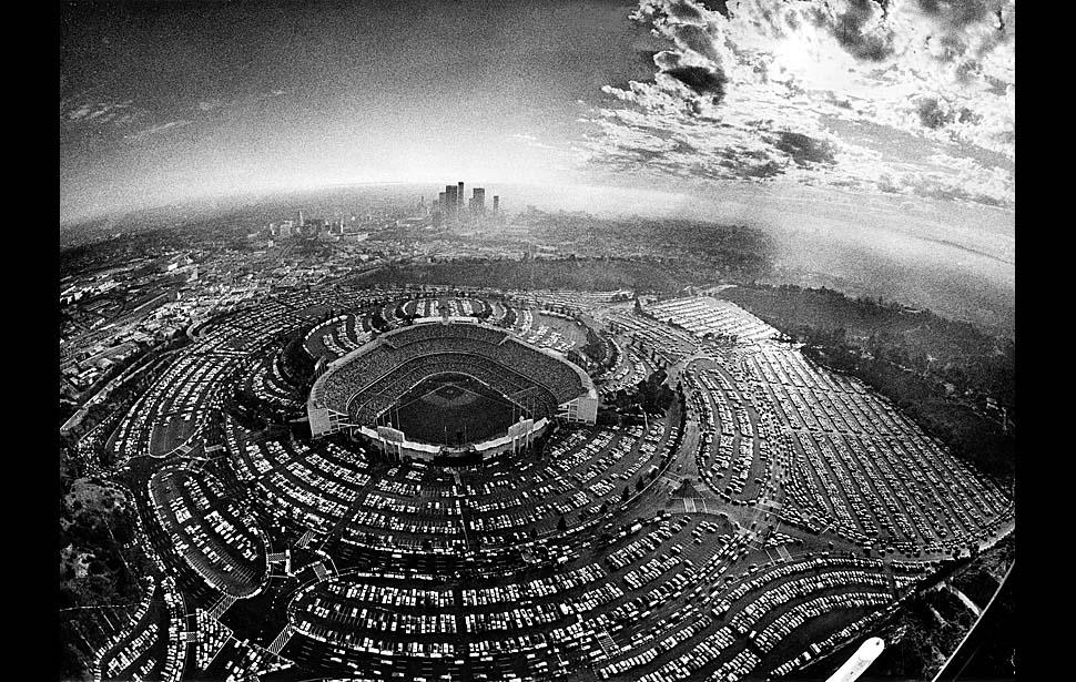 Aerial black-and-white photo of Dodger Stadium surrounded by a full parking lot, with downtown Los Angeles and a cloudy sky visible in the background.