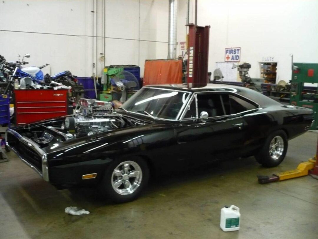 A black classic muscle car with a large exposed engine sits in an auto repair shop, surrounded by tools, red toolboxes, and equipment. A motorcycle is visible in the background.