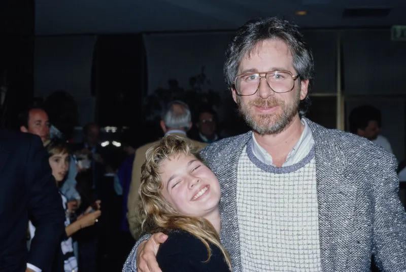 A man with glasses and a beard smiles while hugging a teenage girl with long blonde hair who is grinning with her eyes closed. They appear to be at a social gathering with other people in the background.