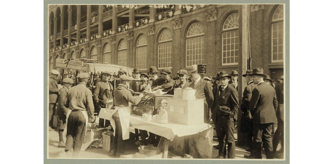 A sepia-toned photo shows men in suits and hats gathered around outdoor food vendors with tables, boxes, and food items in front of a large building with arched windows and a crowd above.