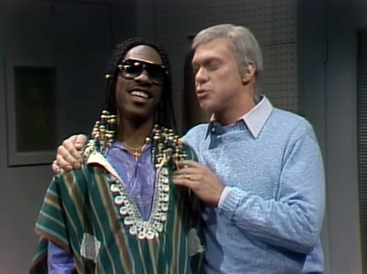 Two men stand close together; one wears sunglasses, a colorful striped poncho, and beaded braids, smiling. The other wears a blue sweater and has a hand on the first man's shoulder, speaking to him.
