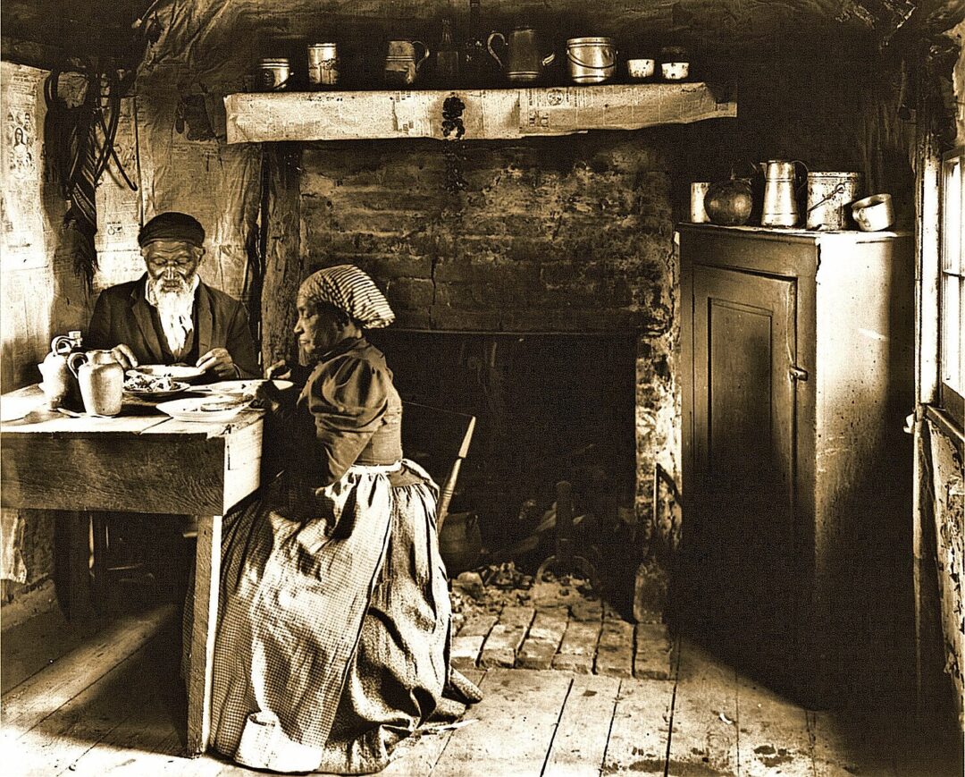 An elderly man and woman sit at a wooden table in a rustic, dimly lit room with a fireplace, simple furnishings, and kitchenware on shelves, evoking a historical, rural setting.