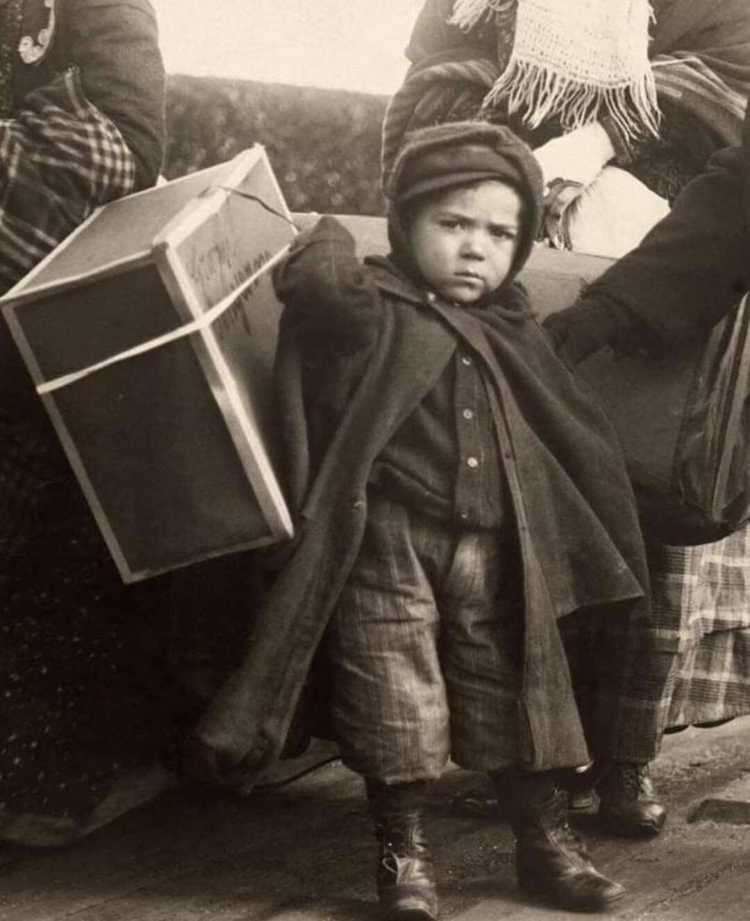 A young child in warm clothing holds a large suitcase, standing among adults at Ellis Island in the early 1900s, representing Italian immigrants arriving in America.