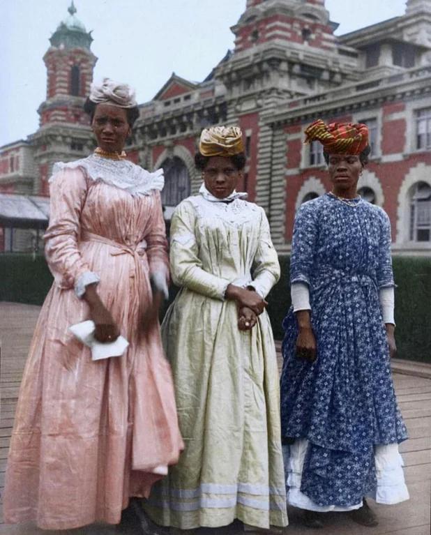 Three women stand outdoors in front of a large historic building, wearing long, colorful 19th-century dresses and headscarves. The building has ornate red brick and white detailing with towers in the background.