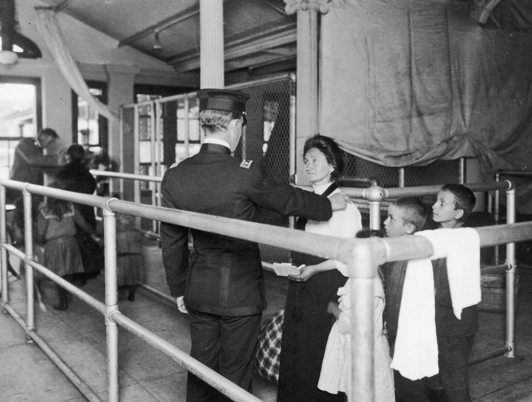 A uniformed officer inspects a woman and three children standing in line in an immigration processing area, likely at Ellis Island, with railings and other people visible in the background.