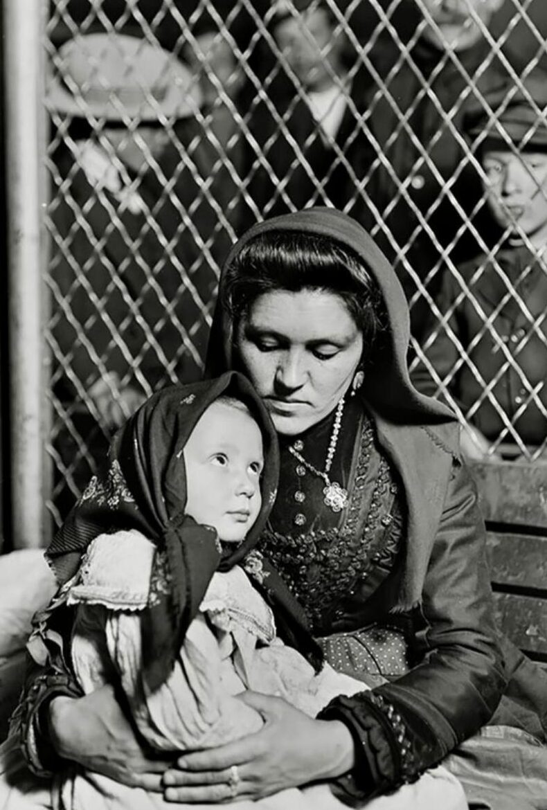 A woman in traditional clothing holds a young child on her lap, both wearing headscarves, sitting in front of a chain-link fence with blurred figures in the background.