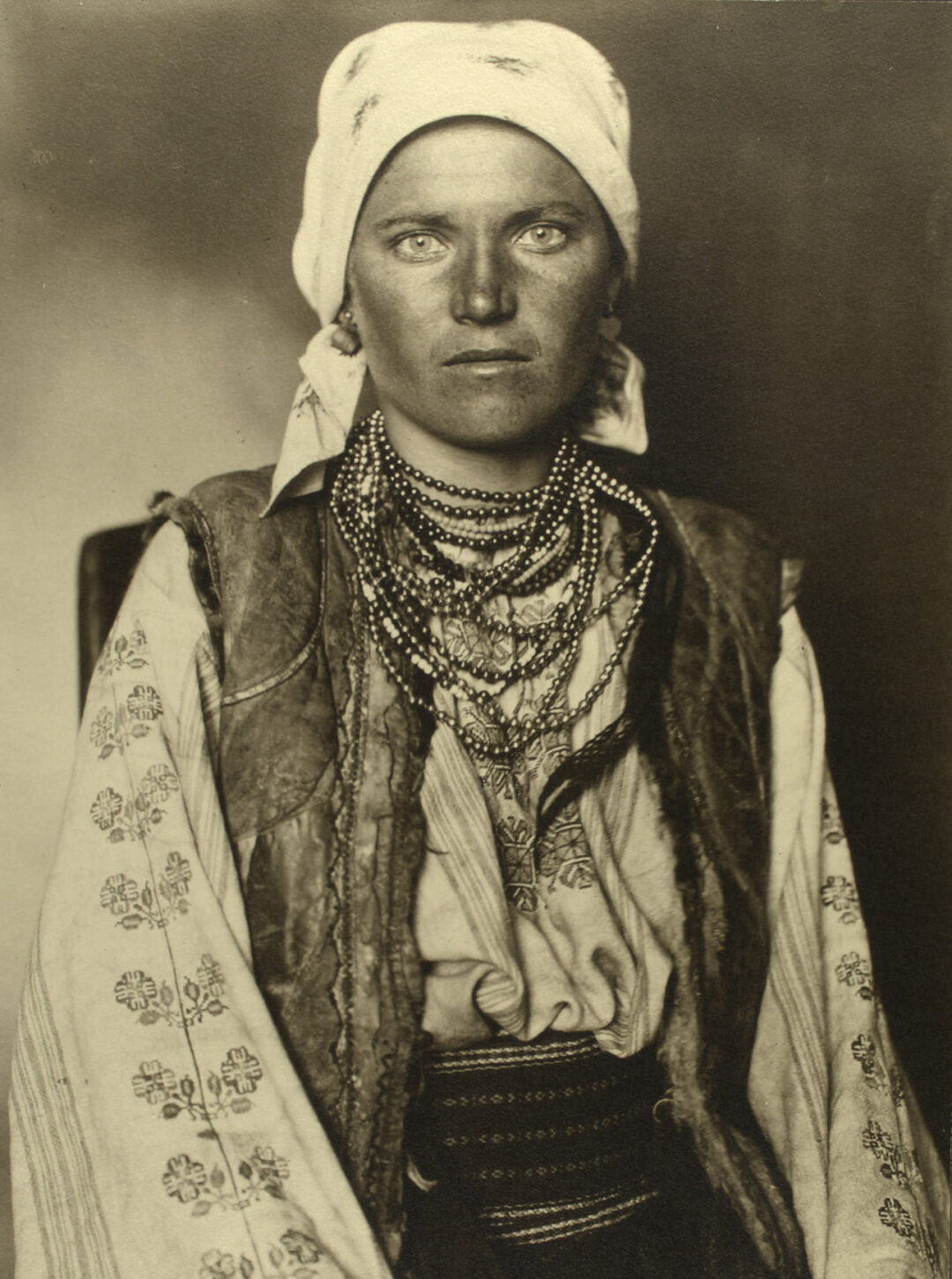 A woman with a serious expression wears traditional Eastern European clothing, including an embroidered blouse, a patterned vest, a headscarf, and multiple beaded necklaces. The photo is sepia-toned and vintage in style.