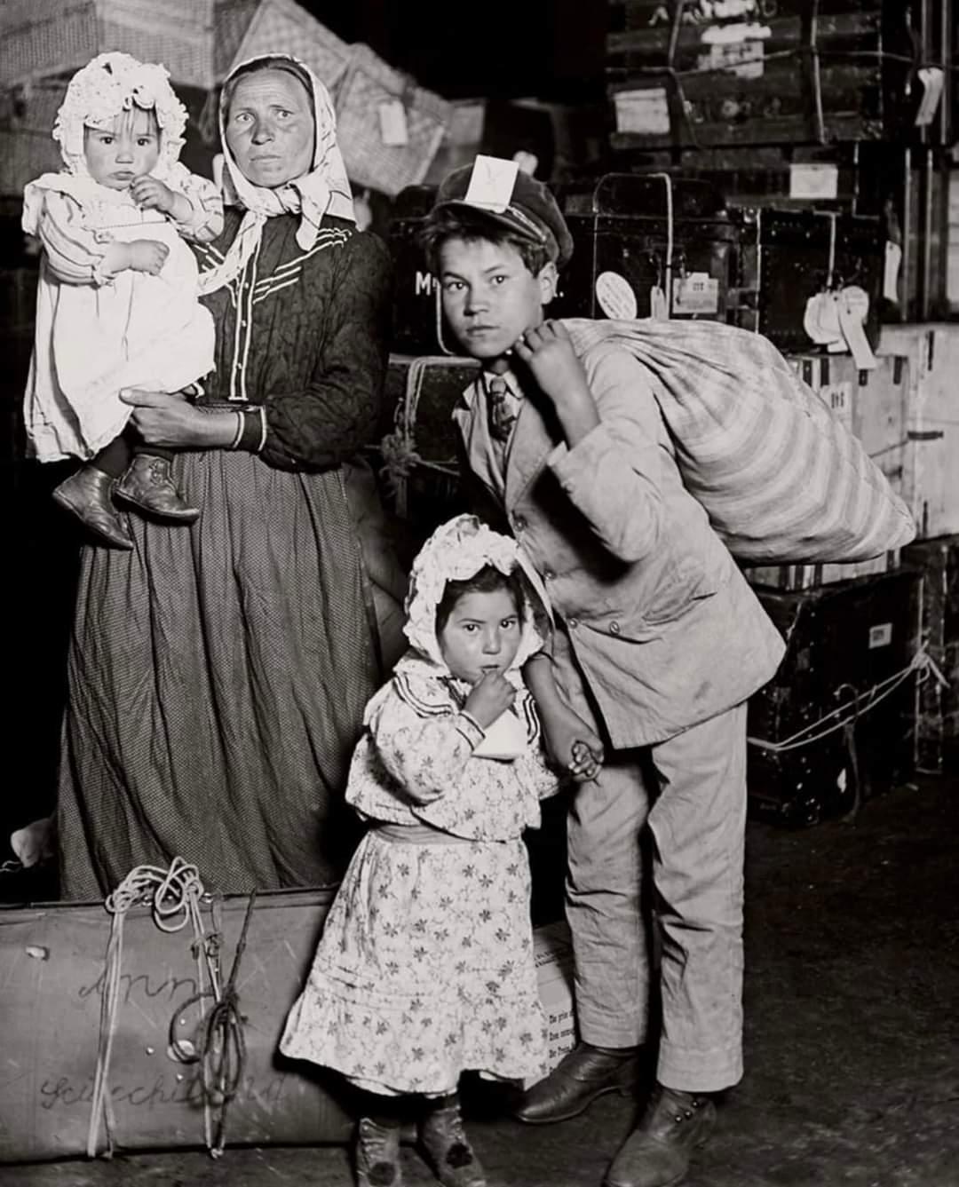 A woman holding a baby stands beside two children, one boy with a sack over his shoulder and one girl in a bonnet, all in old-fashioned clothes, surrounded by luggage in what appears to be an immigration station.