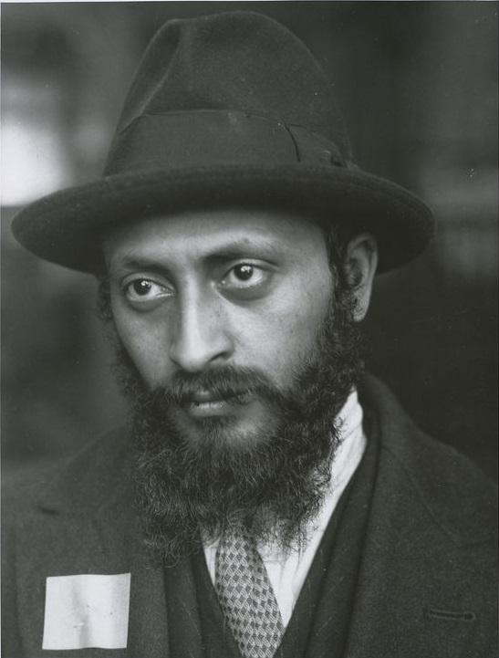 A black-and-white portrait of a man with a full beard and mustache, wearing a dark fedora hat, suit, and tie. He looks slightly to the side with a serious expression.