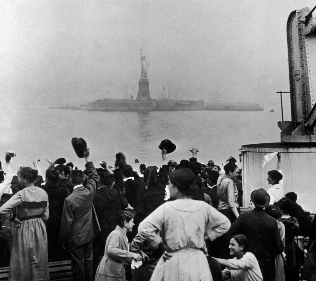 A group of immigrants on a ship’s deck look toward the Statue of Liberty in the distance, some waving hats and hands, as they arrive in New York Harbor on a foggy day.