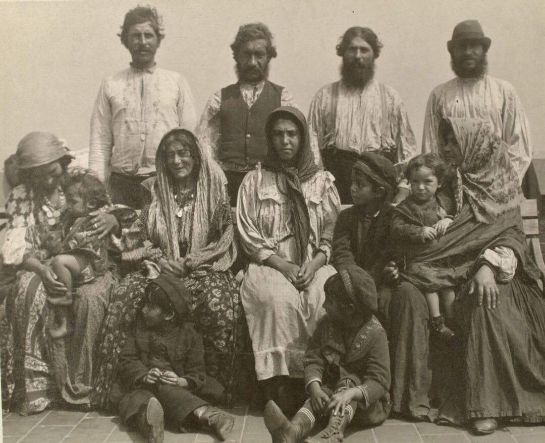 A vintage black-and-white photo of eleven adults and children in traditional clothing, seated and standing in two rows, looking at the camera with serious expressions. The background is plain and light-colored.