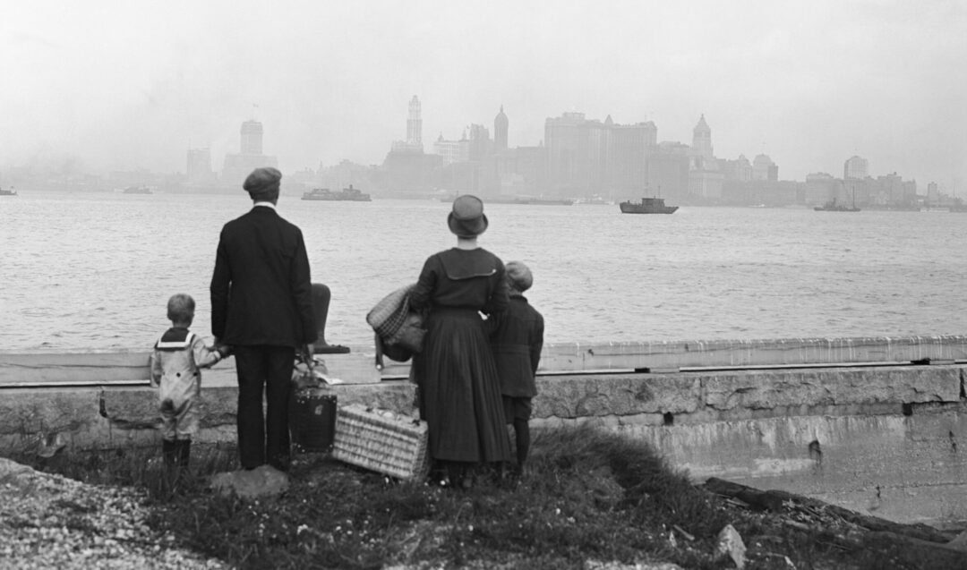A family of five, seen from behind, stands by the water with suitcases, looking toward the distant skyline of New York City, shrouded in mist.