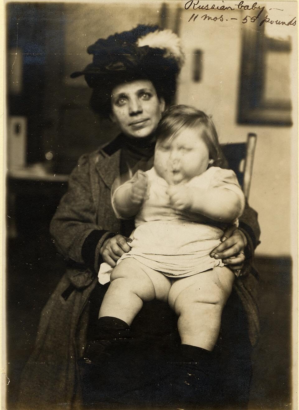 A woman in a hat and coat sits indoors, holding a large baby dressed in light clothing and boots on her lap. Handwritten text at the top right reads: “Russian baby, 11 mos. - 53 1/2 pounds.”