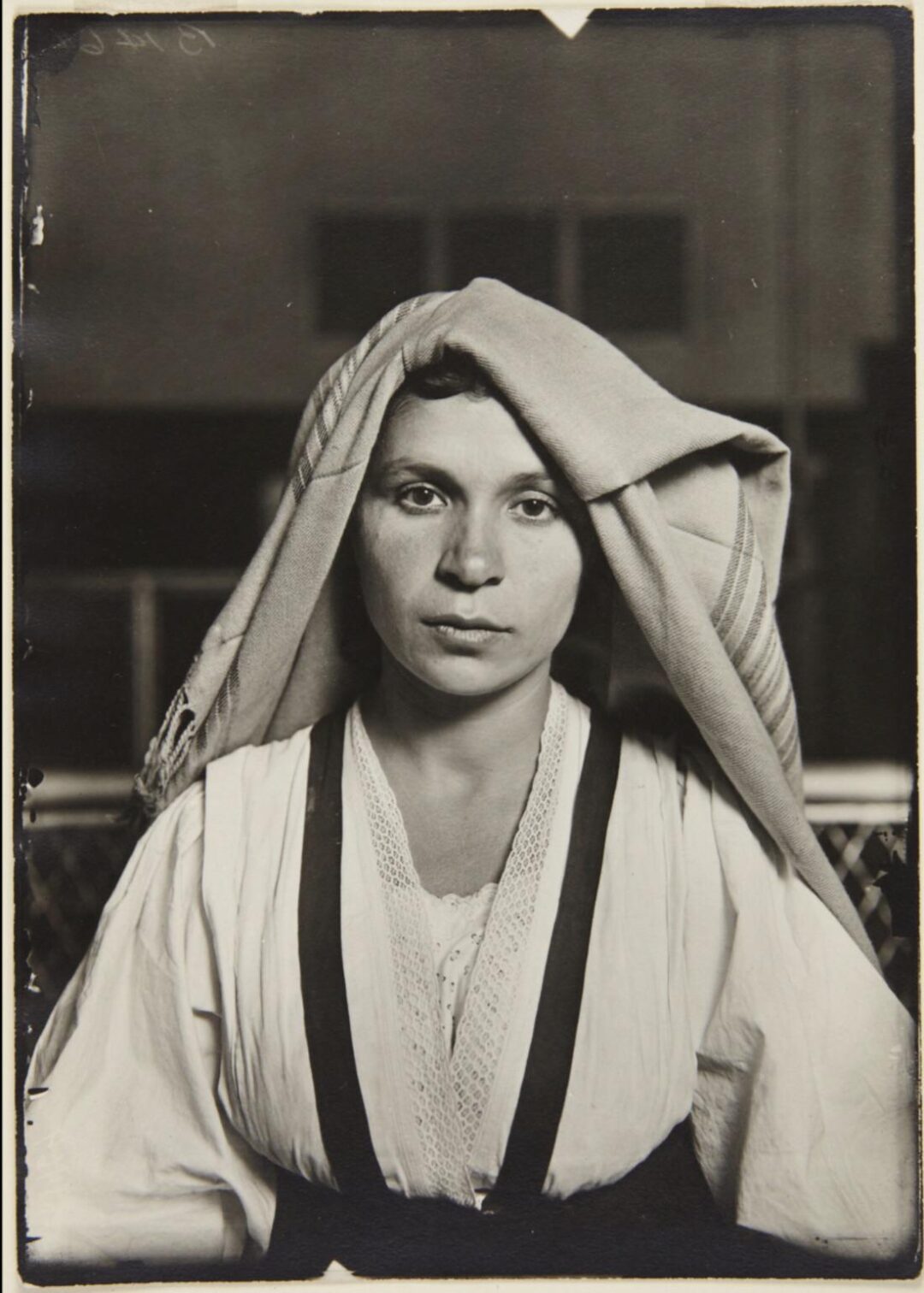 Black and white portrait of a woman wearing traditional clothing and a cloth draped over her head, looking directly at the camera with a neutral expression. The background is out of focus.